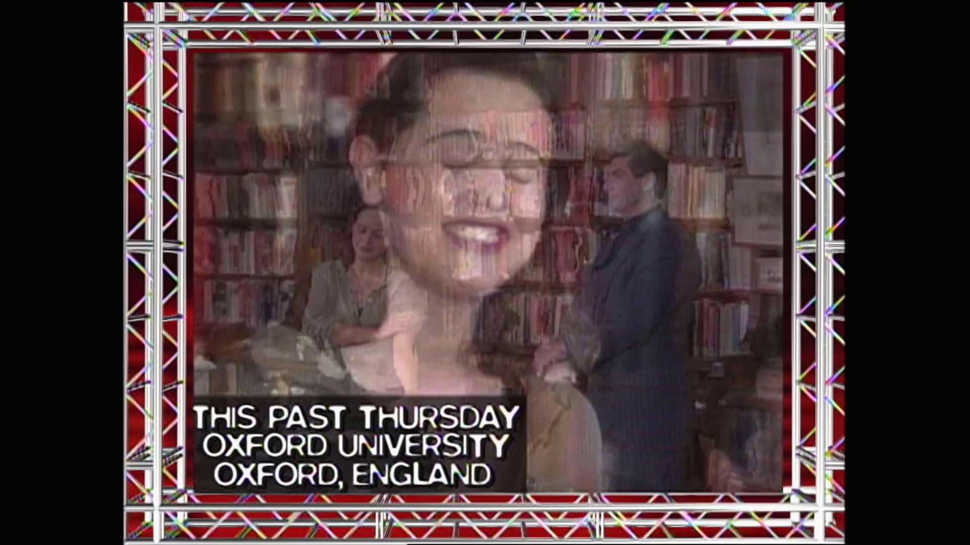 A frame from WWE Raw (December 7, 1998) at 21:45. A large video screen displays a woman smiling in a library setting at Oxford University, with the caption "THIS PAST THURSDAY OXFORD UNIVERSITY OXFORD, ENGLAND" shown below, framed by an ornate red and gold broadcast graphic border.
