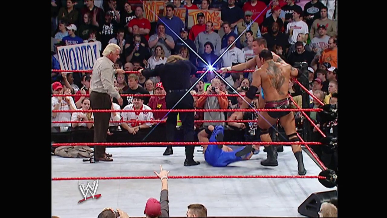 A frame from WWE Raw (February 23, 2004) at 39:44. A WWE Raw match in progress shows a shirtless wrestler being attended to in the ring while an older official stands nearby. The red-roped ring is surrounded by an engaged crowd holding signs, with the WWE logo visible at the bottom of the frame.