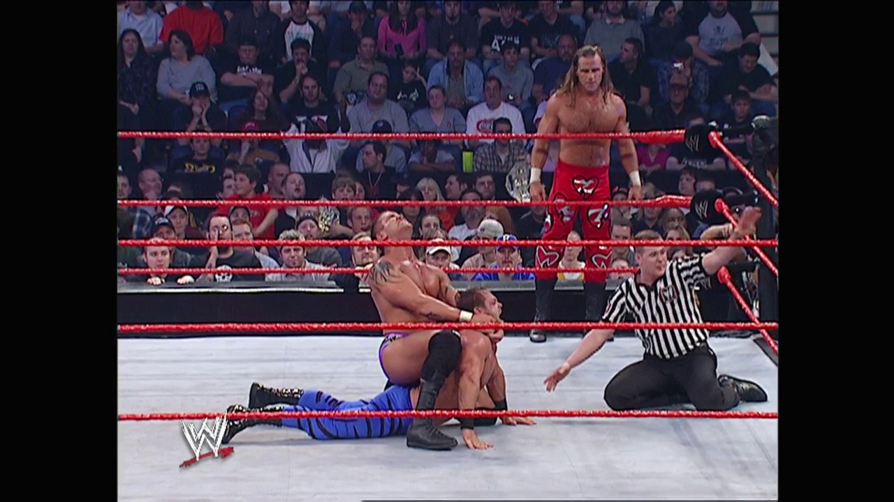 A frame from WWE Raw (March 1, 2004) at 53:25. A WWE wrestler in red attire stands in the ring while another competitor lies on the mat in blue gear. A referee in a striped shirt monitors the action as the crowd watches from the stands, with the red ropes framing the wrestling ring.