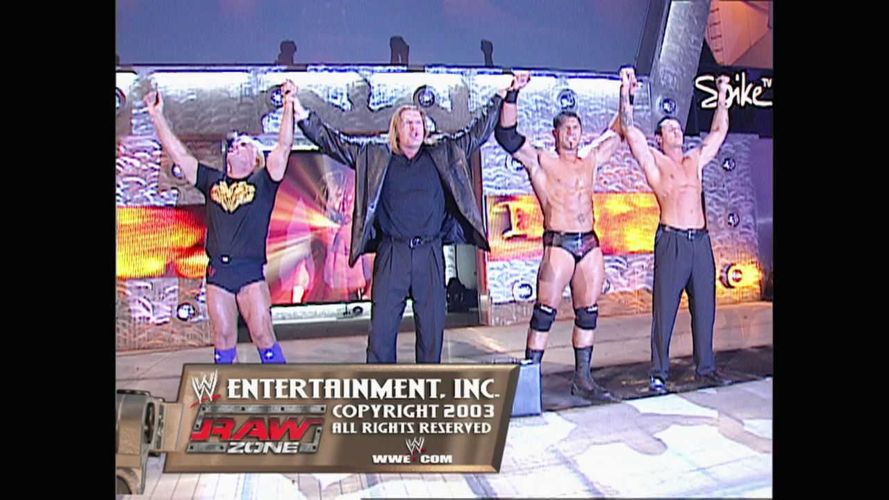 A frame from WWE Raw (October 20, 2003) at 1:33:59. A group of wrestlers stand with raised arms on a WWE Raw stage, with one in a black suit in the center flanked by shirtless competitors. Colorful LED screens and graphics display behind them, with copyright information visible at the bottom from WWE Entertainment, Inc. (2003).