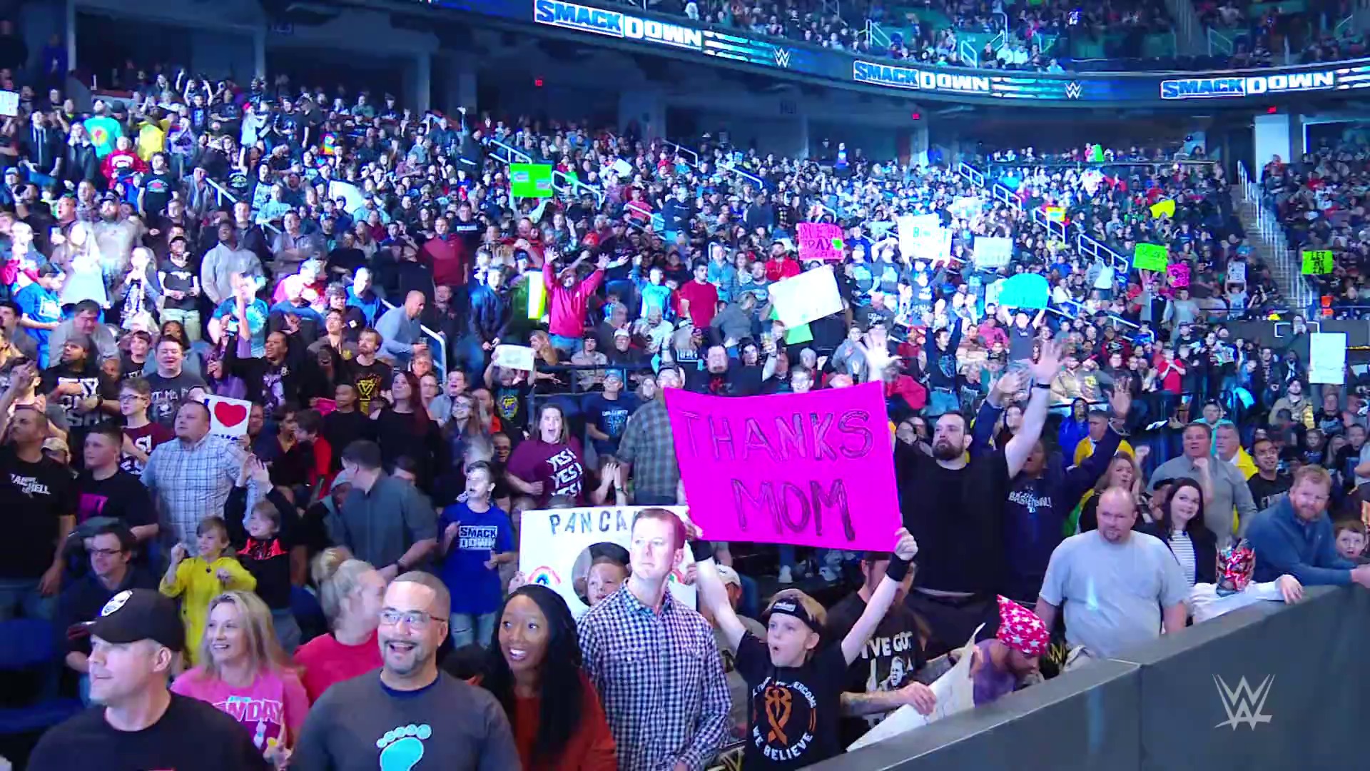 A frame from WWE SmackDown (January 17, 2020) at 14:24. A packed WWE SmackDown crowd enthusiastically holds colorful signs and banners, including a prominent pink "THANKS MOM" sign, while the arena's blue branding and lighting illuminate the venue filled with engaged fans.
