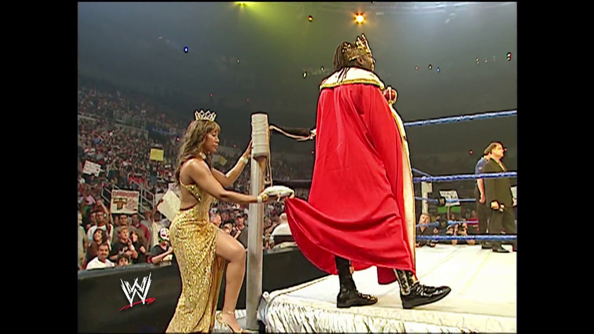 A frame from WWE SmackDown (July 28, 2006) at 1:13:41. A WWE wrestler in a red cape and crown stands on the ring post while another performer in a gold dress presents an award or title belt. A packed crowd fills the arena in the background as the blue-roped ring frames the ceremonial moment.