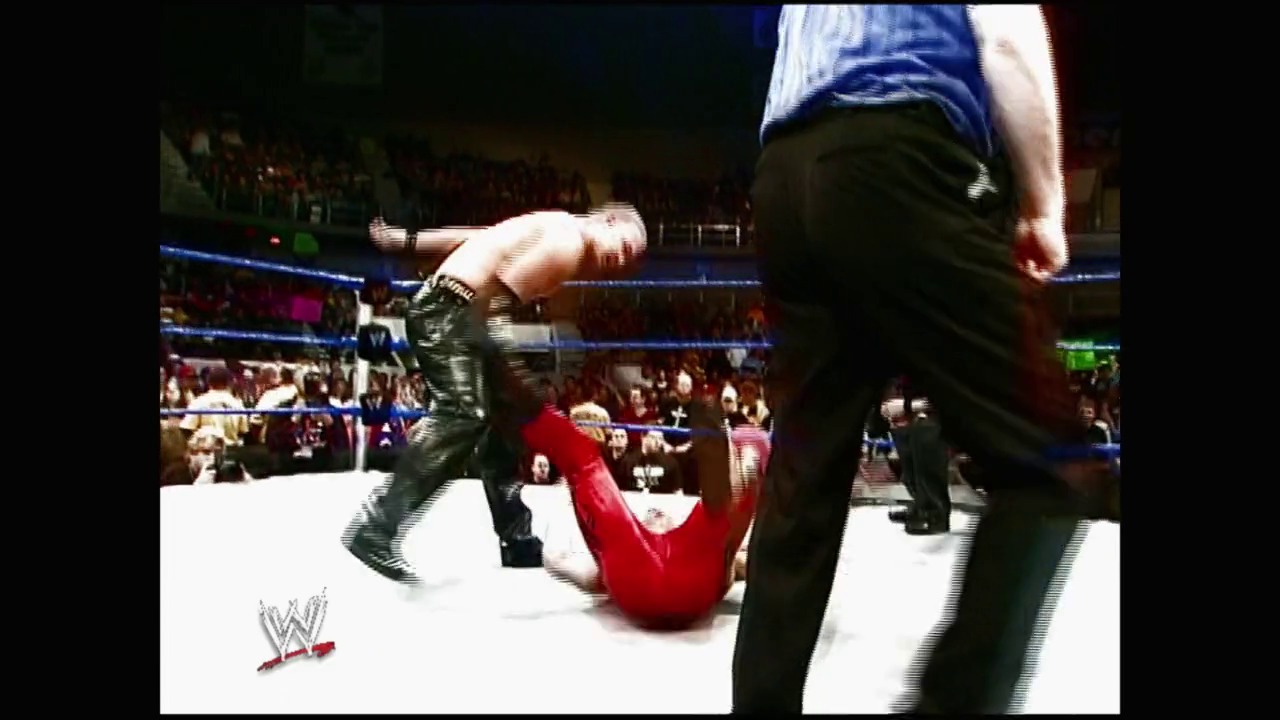 A frame from Royal Rumble (2004) at 23:00. A WWE wrestler in red attire lies on the white canvas while opponents in dark pants stand nearby during a Royal Rumble match, with the crowd visible in the background and the WWE logo displayed in the corner.