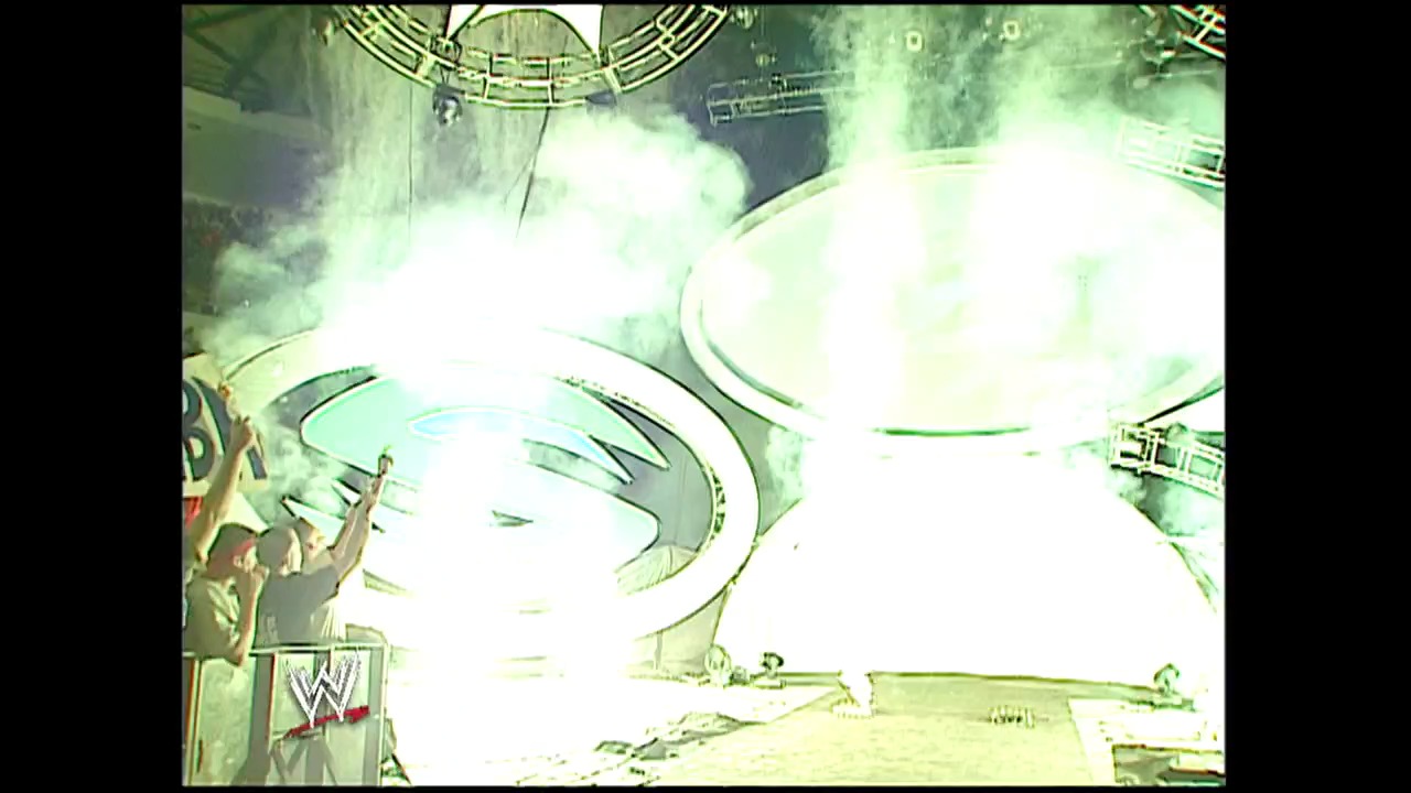 A frame from Summerslam (2002) at 0:17. A wrestling ring is engulfed in bright white smoke and pyrotechnic effects during an intense match at SummerSlam 2002, with the WWE logo visible on the left side and arena structures visible through the haze.