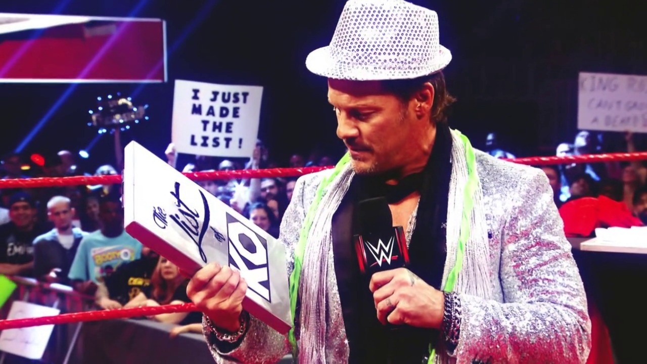 A frame from WrestleMania 33 (2017) at 44:23. A flamboyantly dressed wrestler in a sparkly pink jacket and white fedora holds a WWE sign while speaking into a microphone in the wrestling ring. The crowd and red ropes are visible in the background, with event graphics and signage displayed around the arena.
