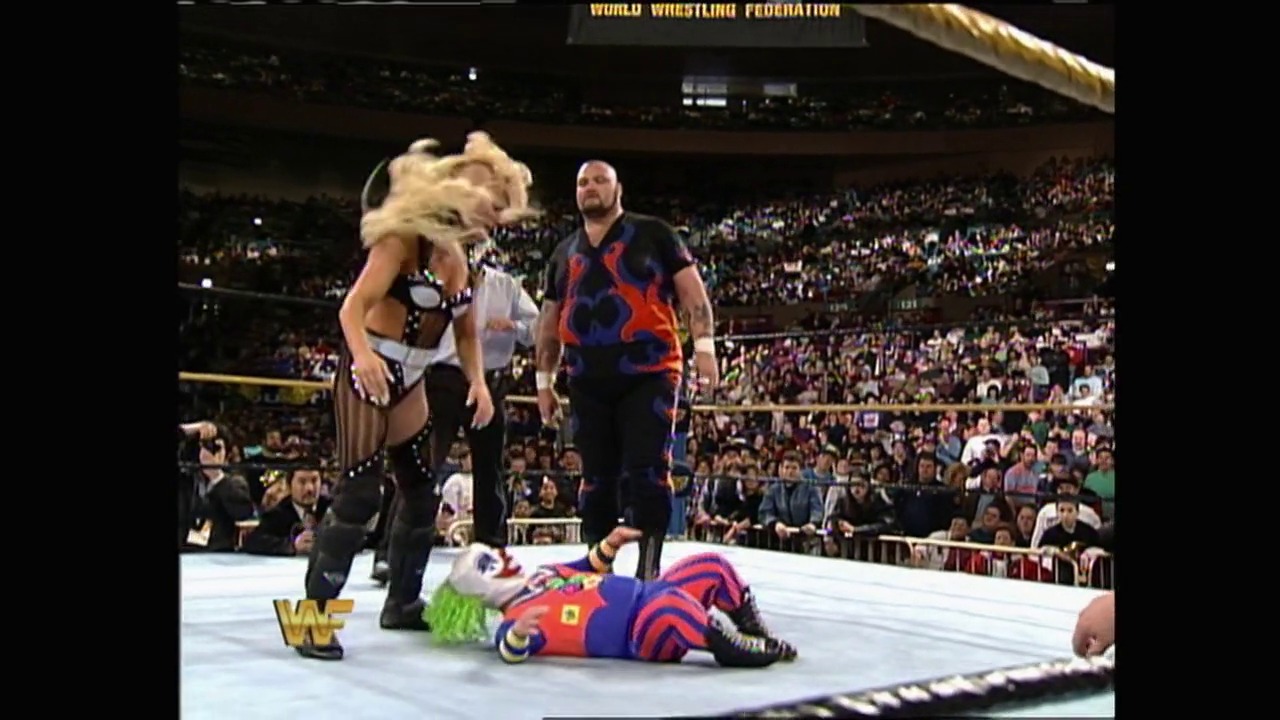 A frame from WrestleMania X (1994) at 47:02. A WWF wrestler in colorful red and blue attire lies on the mat while two other wrestlers stand nearby in a packed arena. The crowd fills the stadium seats in the background, with the WWF logo visible in the corner of the broadcast.