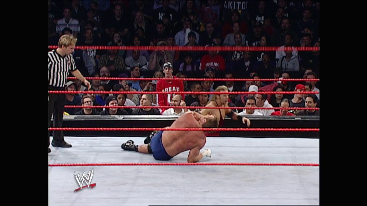 A frame from WWE Raw (February 10, 2003) at 13:37. A wrestler lies on the canvas in a WWE Raw ring surrounded by red ropes, while a referee in a striped shirt stands nearby. The crowd watches from the stands in the background, with the WWE logo visible in the corner of the frame.