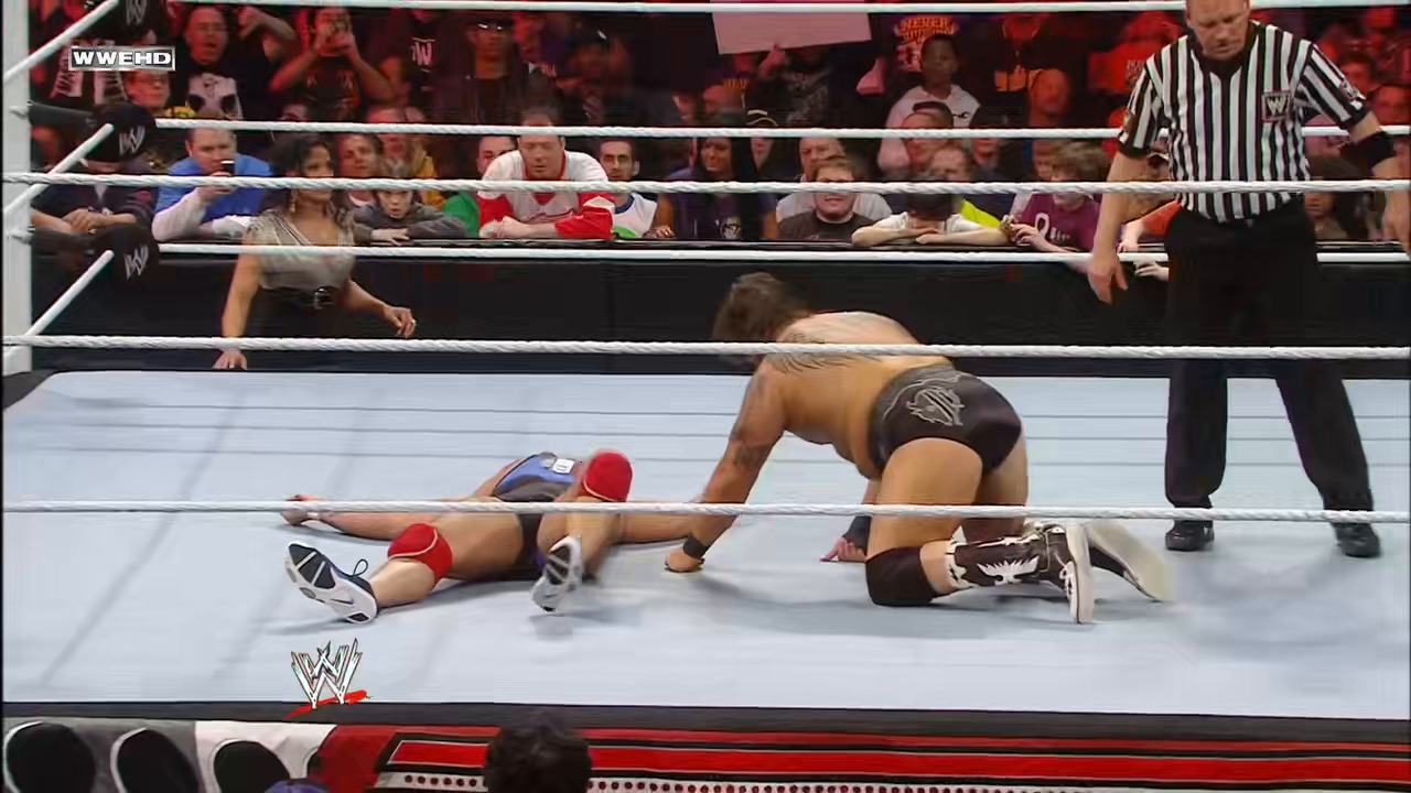 A frame from WWE Raw (January 24, 2011) at 1:16:43. Two wrestlers are down on the WWE Raw mat during a match, with one pinned and the referee in position to count. The crowd watches intently from the stands while the referee officiates the action in the ring.