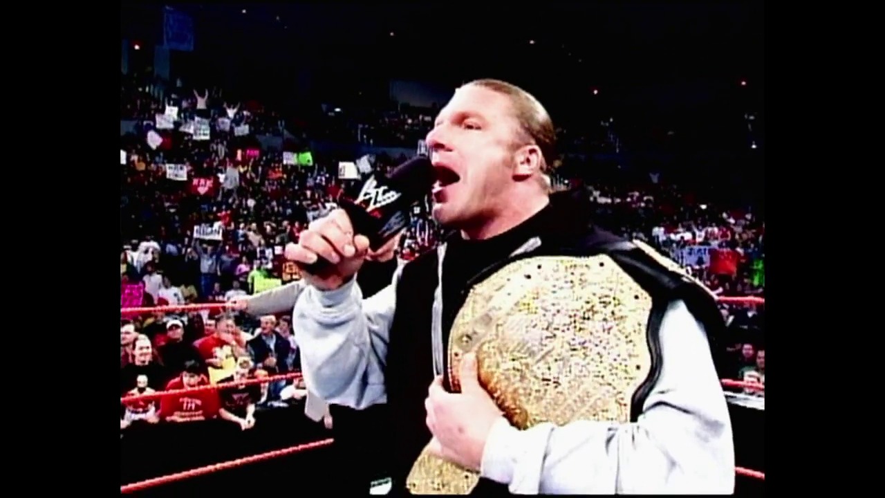 A frame from WWE Raw (January 6, 2003) at 0:39. A wrestler holds a microphone and championship belt while addressing a packed arena during a WWE Raw event, with the enthusiastic crowd visible in the background under arena lighting.