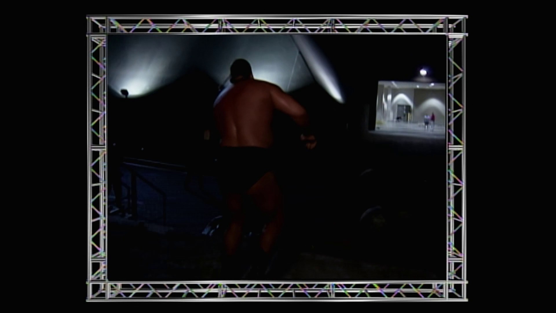 A frame from WWE Raw (June 21, 1999) at 1:13:13. A shirtless wrestler stands in the darkened ring under dramatic overhead lighting during a WWE Raw broadcast. The scene is framed by silver metal trussing, with a monitor visible in the upper right corner showing additional event footage.