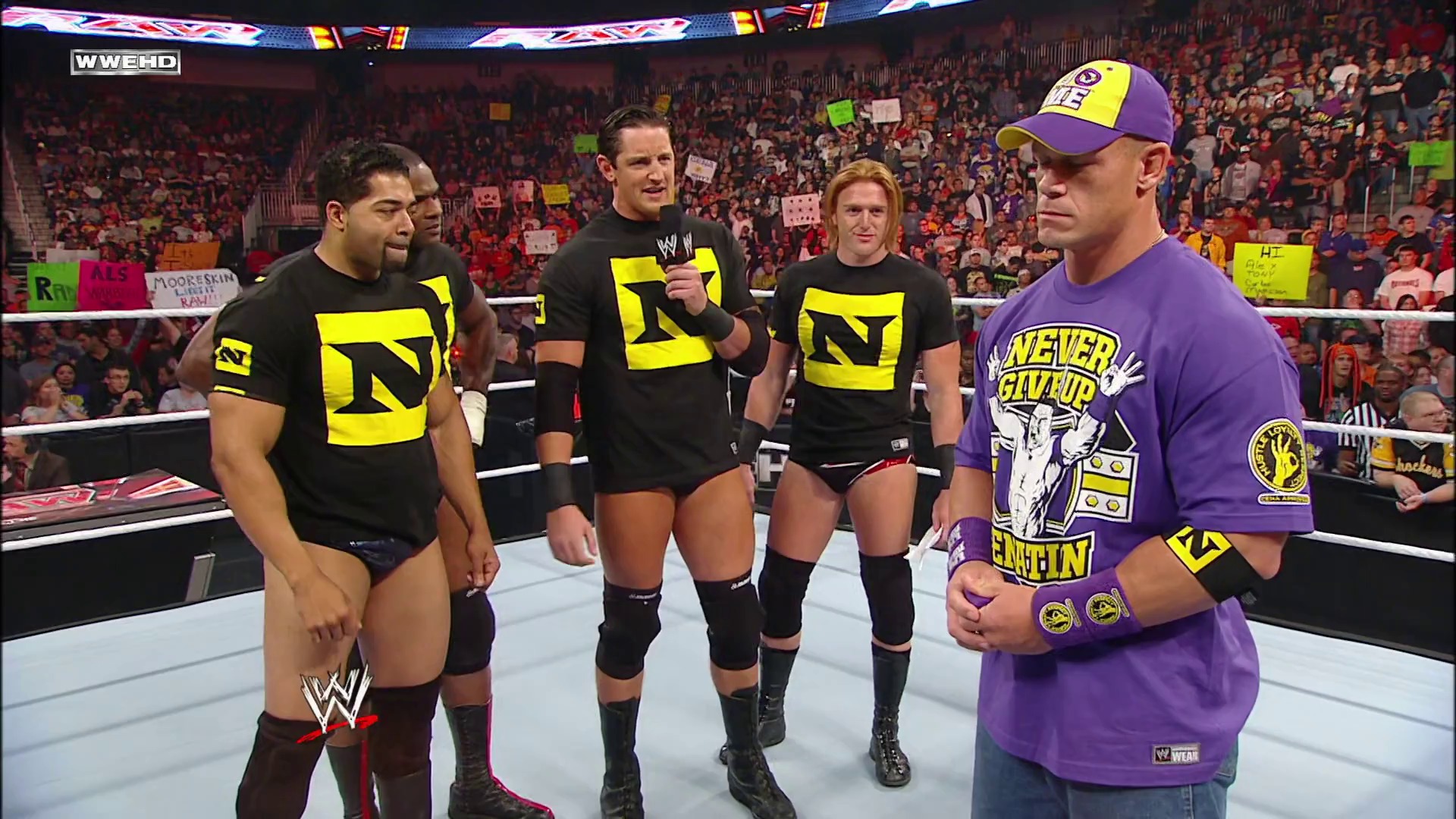 A frame from WWE Raw (October 4, 2010) at 13:09. # WWE Raw Scene

A WWE wrestler in purple attire and cap confronts three opponents wearing black and yellow shirts in the ring, with a packed arena crowd visible in the background and the white WWE ring ropes in the foreground.