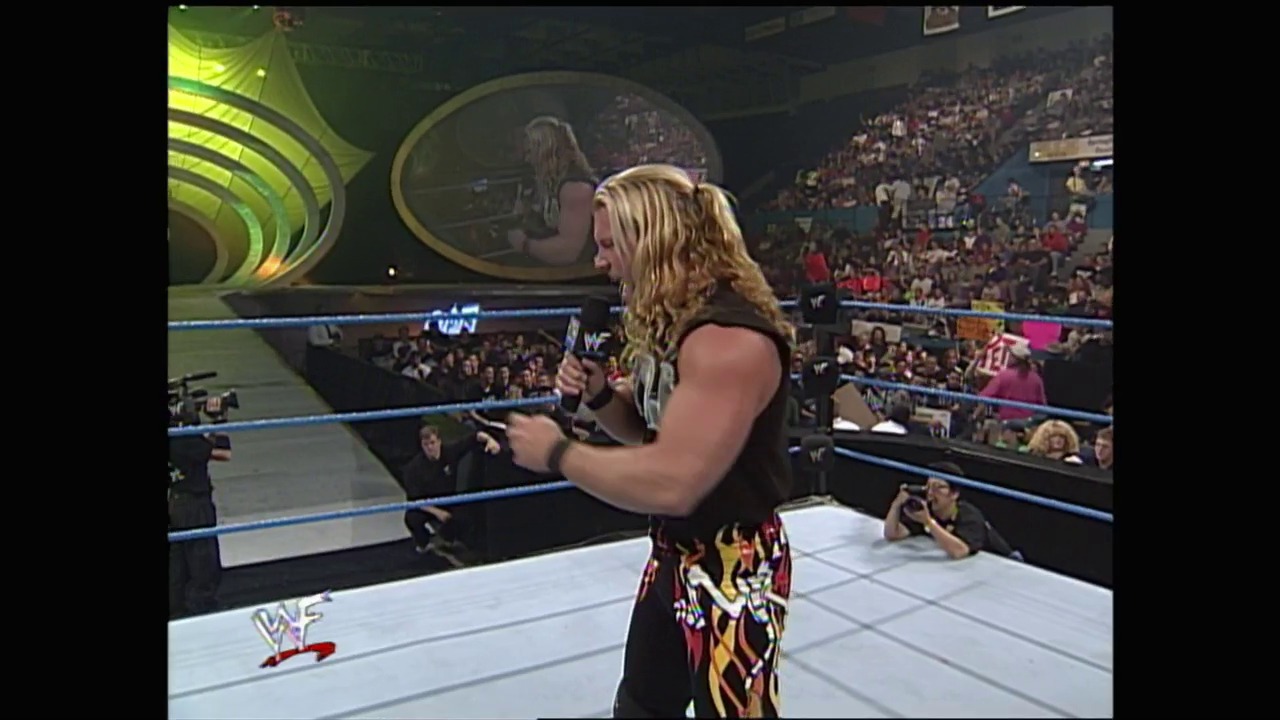 A frame from WWE SmackDown (October 28, 1999) at 25:00. A blonde wrestler stands in the WWF ring holding a microphone, addressing the packed arena crowd. The wrestler wears dark attire with flame designs, positioned near the blue ropes with the WWF logo visible on the mat.
