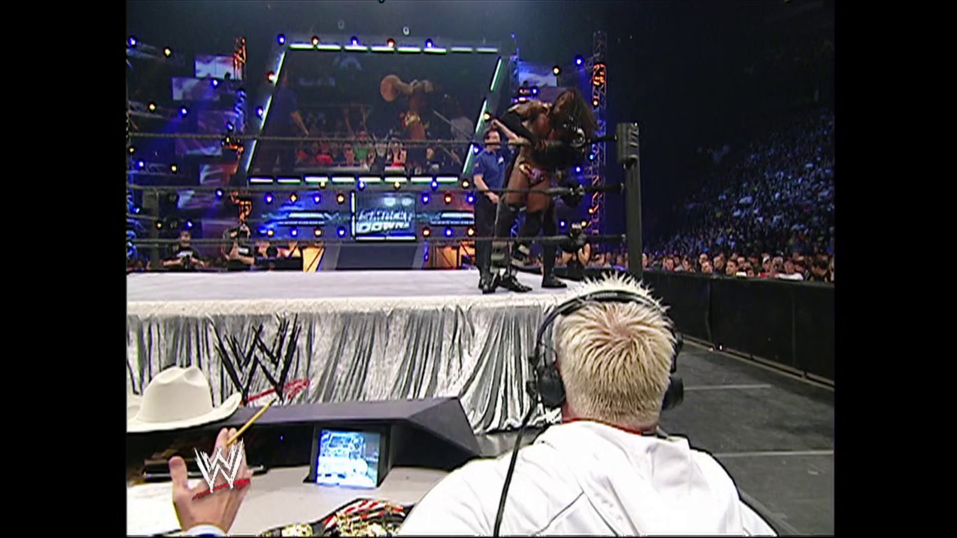 A frame from WWE SmackDown (September 22, 2006) at 1:15:29. A WWE SmackDown broadcast from the commentator's booth shows a wrestler performing in the illuminated ring while the crowd fills the arena. Blue and gold lighting frames the wrestling ring with its distinctive WWE branding visible on the apron.