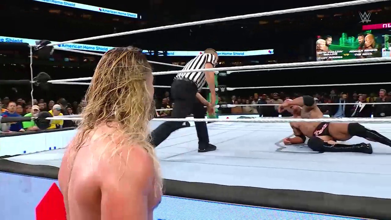 A frame from WWE WrestleMania 40 Saturday (2024) at 3:40:15. A WWE wrestling match in progress at WrestleMania 40, with a shirtless wrestler in the foreground watching from ringside as a referee tends to an opponent on the mat. The illuminated white ring, crowd, and "American Home Shield" sponsorship boards are visible in the arena.