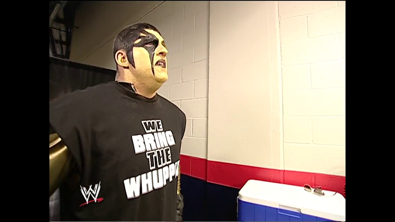 A frame from King of the Ring (2002) at 1:54:56. A wrestler with distinctive face paint stands in a backstage area wearing a black "We Bring the Whoop" WWE t-shirt, positioned in front of a cooler near a red and blue striped wall.