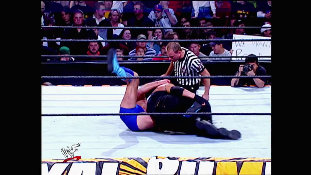 A frame from Royal Rumble (2002) at 46:35. A WWE wrestler in blue trunks lies on the mat during a Royal Rumble 2002 match, while a referee in striped shirt stands nearby. The crowd watches intently from the stands behind the ring ropes.