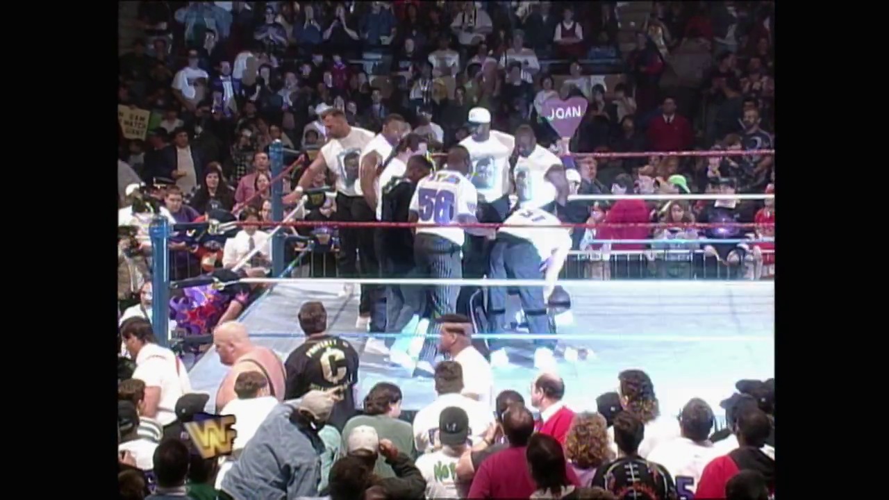A frame from WrestleMania XI (1995) at 2:24:32. A wrestling match is in progress at WrestleMania XI, with multiple wrestlers in the ring wearing white and blue attire, while spectators crowd around the perimeter and filled stands cheer from above.