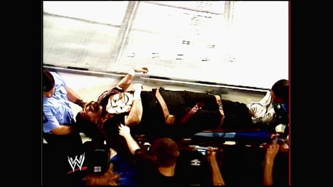 A frame from WWE Raw (February 23, 2004) at 1:05:14. A WWE wrestler is being lifted and thrown during a match, captured from an overhead angle. The scene shows intense action with multiple people involved, the ring ropes visible, and the WWE logo in the bottom left corner.