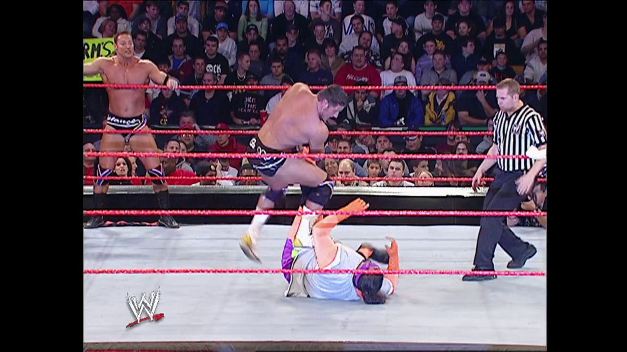 A frame from WWE Raw (November 10, 2003) at 27:27. A WWE wrestler lies on the mat after being knocked down, while another shirtless competitor stands over him. A referee in a striped shirt watches nearby as the crowd fills the arena seats behind the red-roped ring.