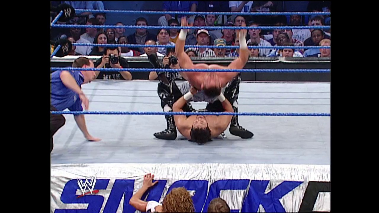 A frame from WWE Raw (October 24, 2002) at 24:09. A wrestler in black leather attire lies on their back in a WWE ring, arms raised in victory, while another competitor in blue stands nearby. The crowd fills the stands behind the blue-roped ring, with the WWE SmackDown logo visible at the bottom of the frame.