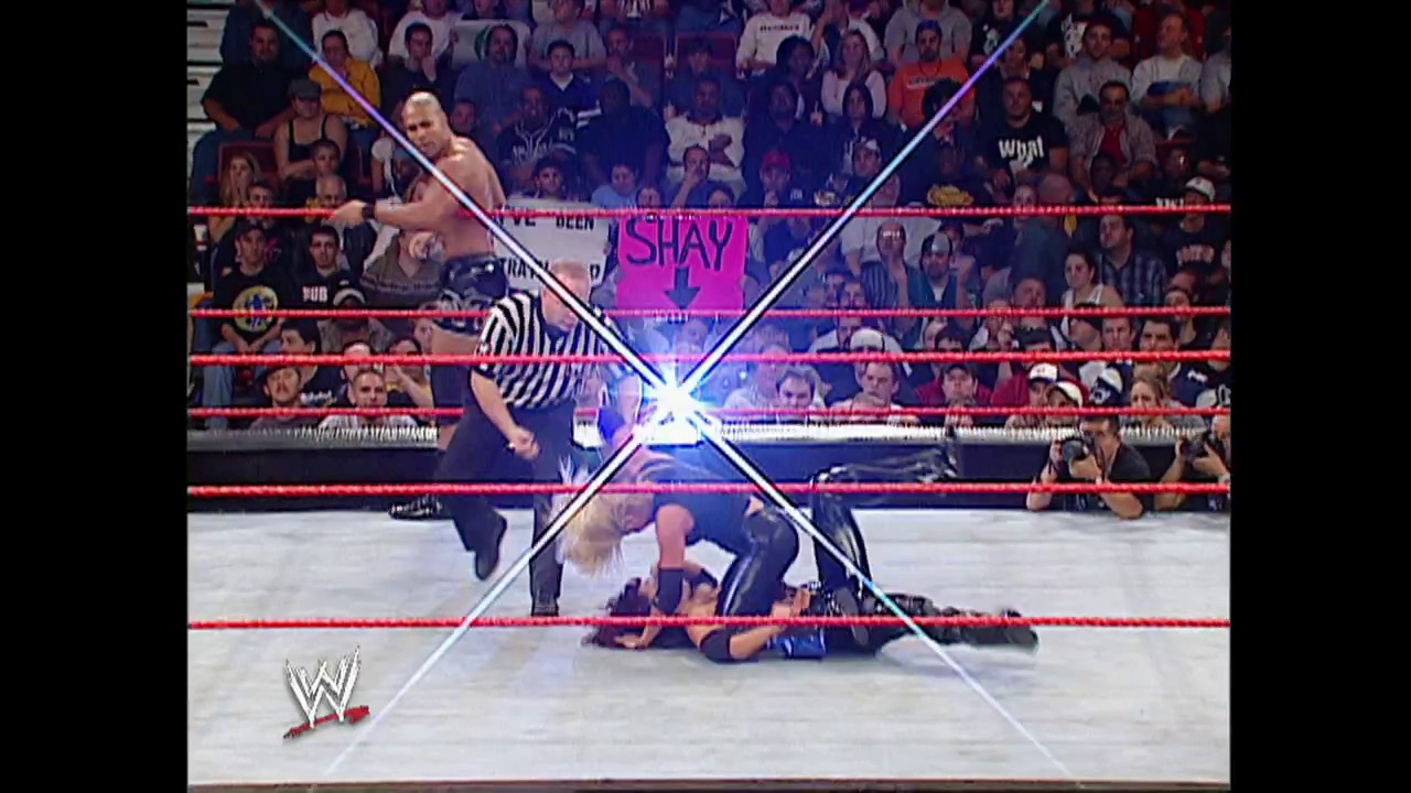 A frame from WWE Raw (October 6, 2003) at 1:19:03. A WWE wrestler in striped referee attire stands in the ring while another performer lies on the mat below. The crowd fills the arena background, with a pink "SHAY" graphic visible above the ring and the WWE logo displayed in the corner.