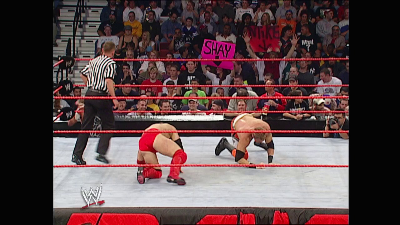 A frame from WWE Raw (October 6, 2003) at 53:18. A WWE Raw match in progress featuring two wrestlers in the red-roped ring, with a referee in striped shirt overseeing the action. The crowd fills the arena stands in the background, and a "SHAY" sign is visible among the spectators.