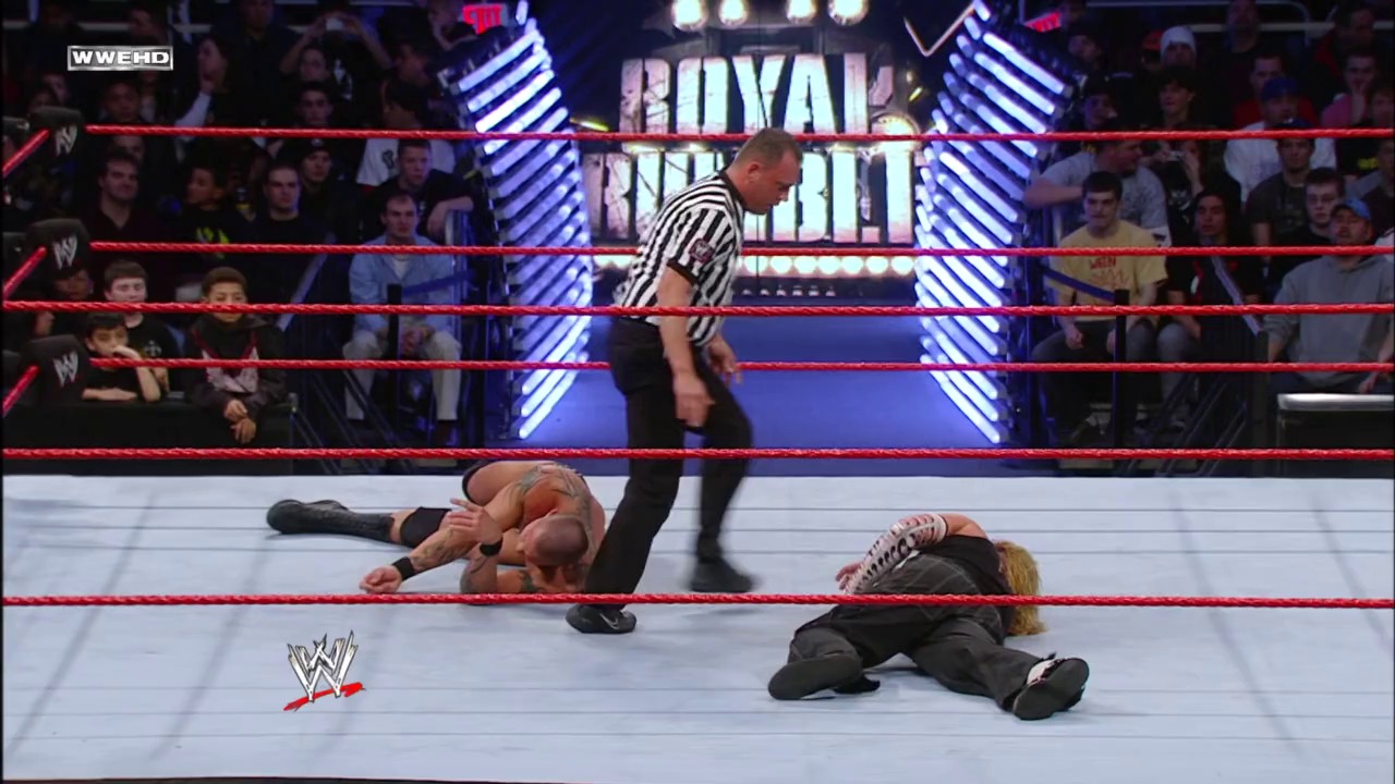 A frame from WWE Royal Rumble (2008) at 1:35:13. A WWE referee stands over two wrestlers lying on the mat during the 2008 Royal Rumble event, with the illuminated "Royal Rumble" signage visible above the red-roped ring and a packed crowd surrounding the arena.