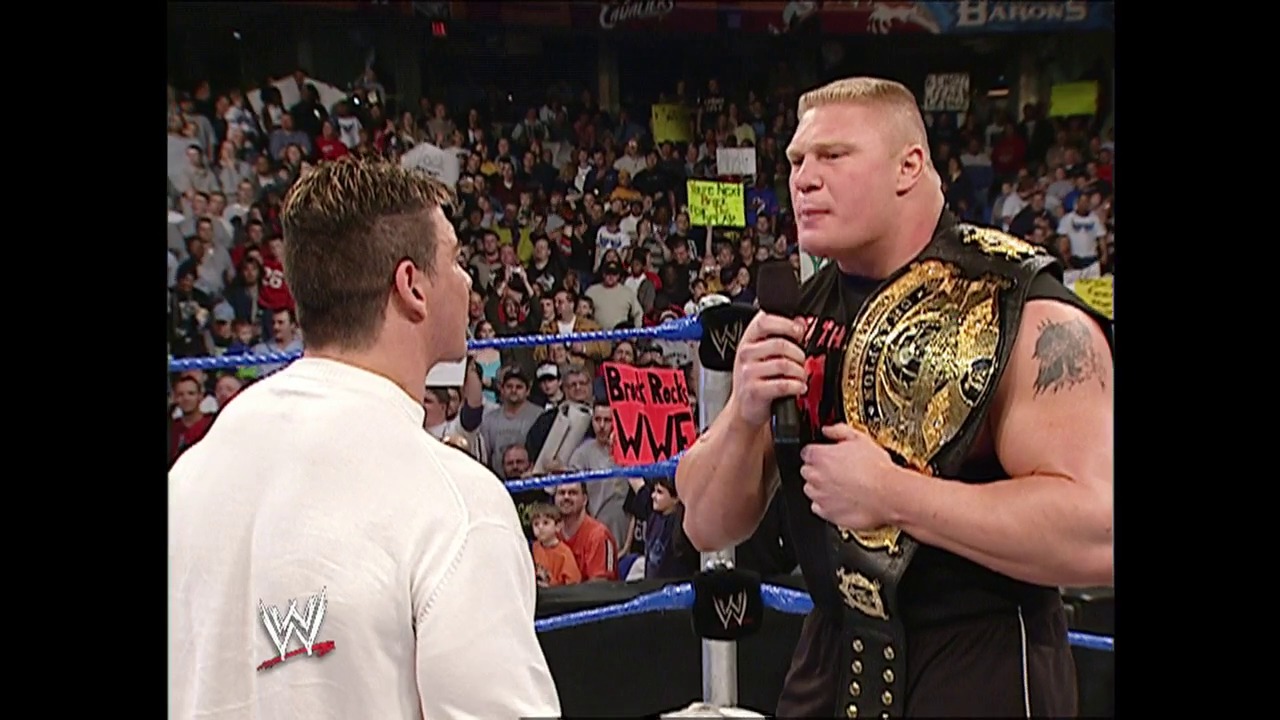 A frame from WWE SmackDown (February 5, 2004) at 37:26. A WWE wrestler in a black outfit with a gold championship belt holds a microphone while speaking to another wrestler in a white shirt inside the ring. A packed crowd fills the arena in the background.