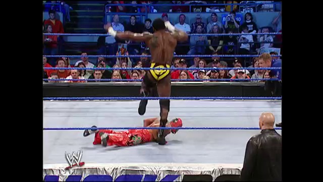 A frame from WWE SmackDown (March 11, 2005) at 1:11:30. A wrestler in black trunks stands over a fallen opponent in red attire on the WWE SmackDown ring, with the crowd visible in the background and a referee present ringside.
