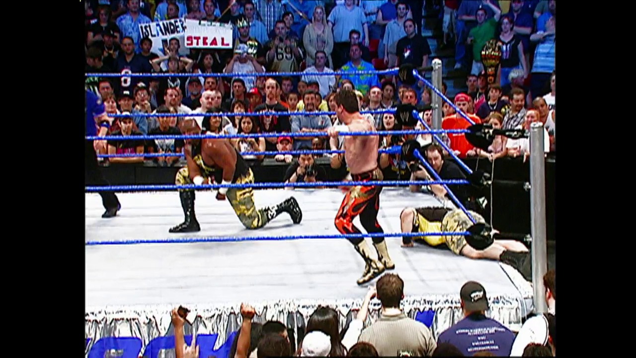 A frame from WWE SmackDown (May 27, 2004) at 25:01. A WWE wrestler in red and gold attire stands in the ring while opponents in black and gold lie on the mat. The crowd fills the arena stands behind the blue-roped ring, with visible signage and ringside personnel.