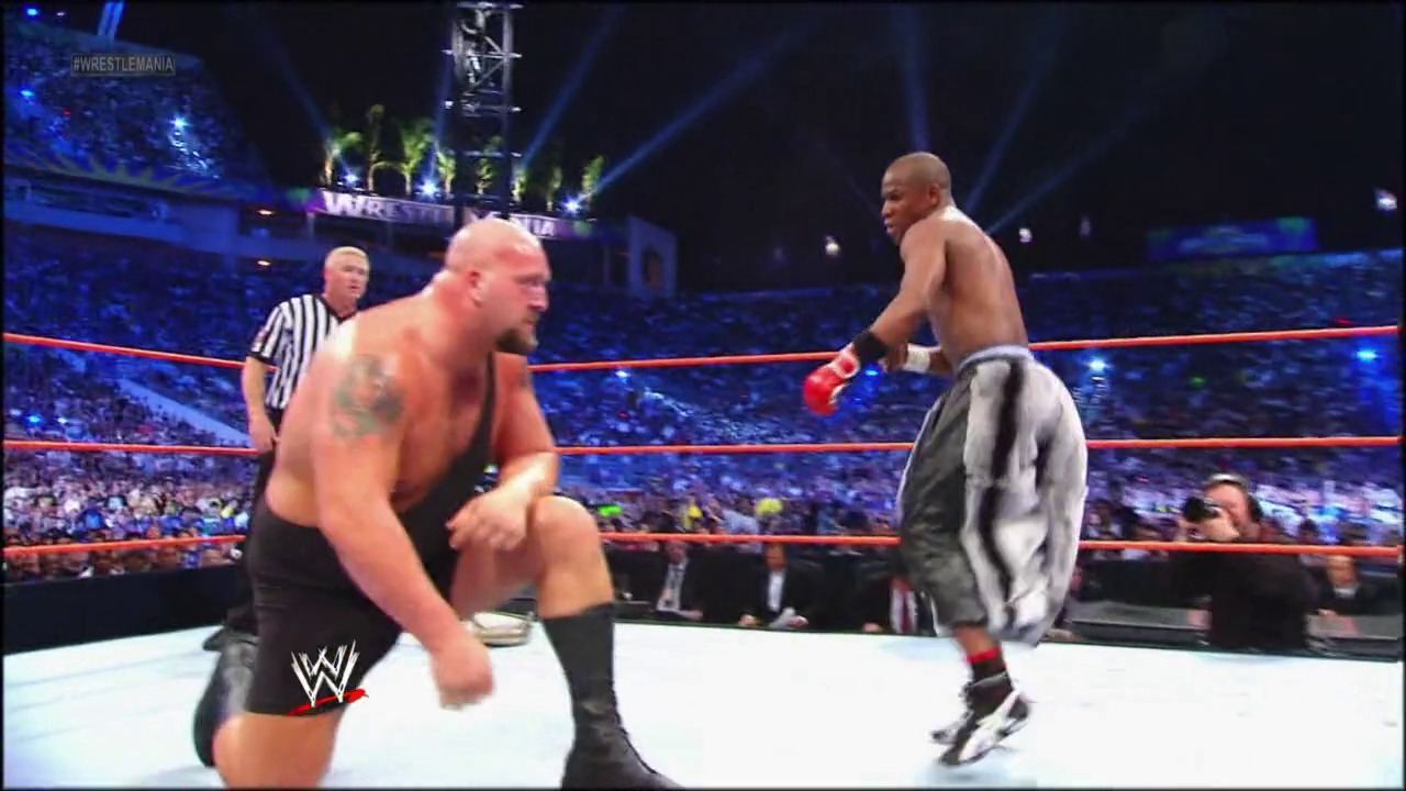 A frame from WrestleMania XXVIII (2012) at 33:51. Two shirtless wrestlers face off in a WWE ring during WrestleMania XXVIII, with a referee nearby and a massive crowd visible in the blue-lit arena background. The wrestler on the left is in a fighting stance while his opponent approaches from the right.