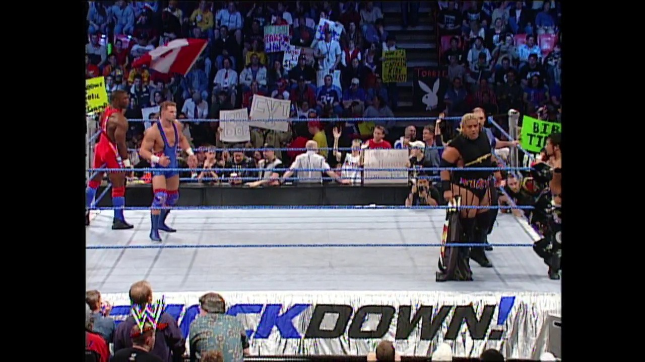 A frame from WWE SmackDown (May 8, 2003) at 1:00:37. A WWE SmackDown match in progress featuring wrestlers in the ring with the blue ropes visible, a large crowd in the background holding signs, and the "SmackDown" logo displayed at the bottom of the frame.