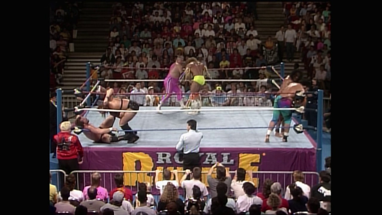 A frame from Royal Rumble (1990) at 2:19:21. A wrestler lies on the mat while others stand in the ring during a Royal Rumble match, with the referee positioned ringside and a large crowd visible in the bleachers above the purple-skirted ring adorned with the event's branding.