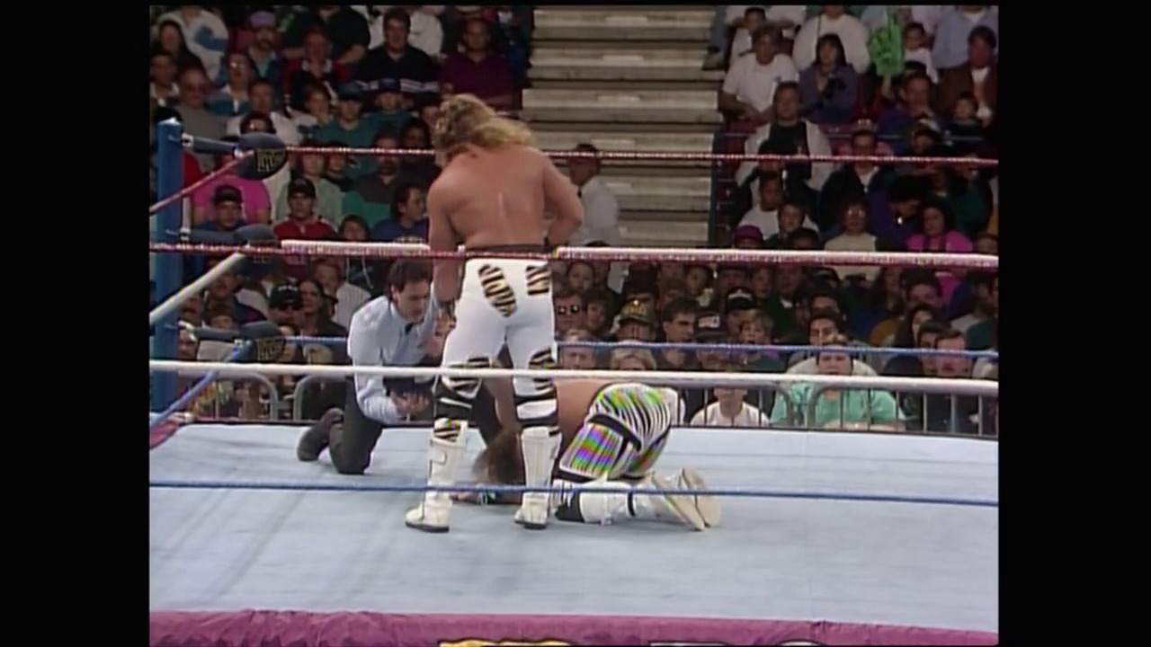 A frame from Royal Rumble (1993) at 26:36. A wrestler in white and gold attire is on the mat in a WWF ring, with an opponent standing nearby. The crowd fills the arena in the background, separated by the distinctive red ropes of the wrestling ring.