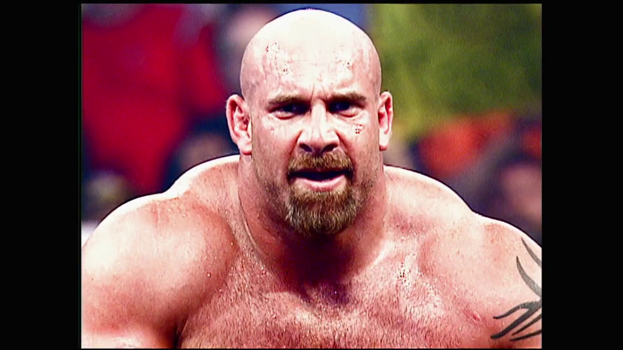 A frame from WWE Bad Blood (2003) at 0:45. A muscular, bald wrestler with a goatee and visible tattoo is shown in close-up during a WWE event, appearing intense during competition. The blurred arena background with red and green lighting suggests an active wrestling match with an engaged crowd.