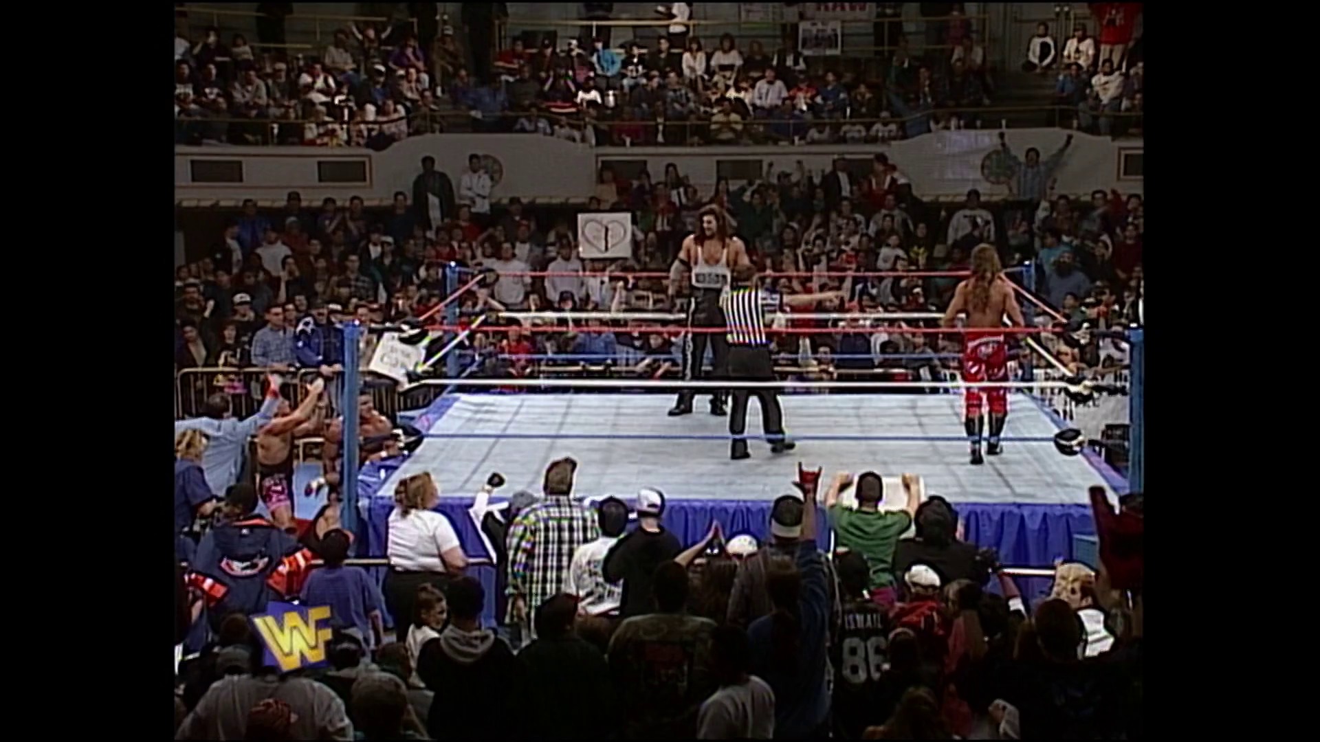 A frame from WWE Raw (February 5, 1996) at 13:09. A WWE wrestling match is in progress with a referee in the ring and wrestlers positioned on the canvas. The arena is packed with enthusiastic fans in the stands, with the blue and red WWE ring ropes visible and the crowd barrier separating spectators from the action.