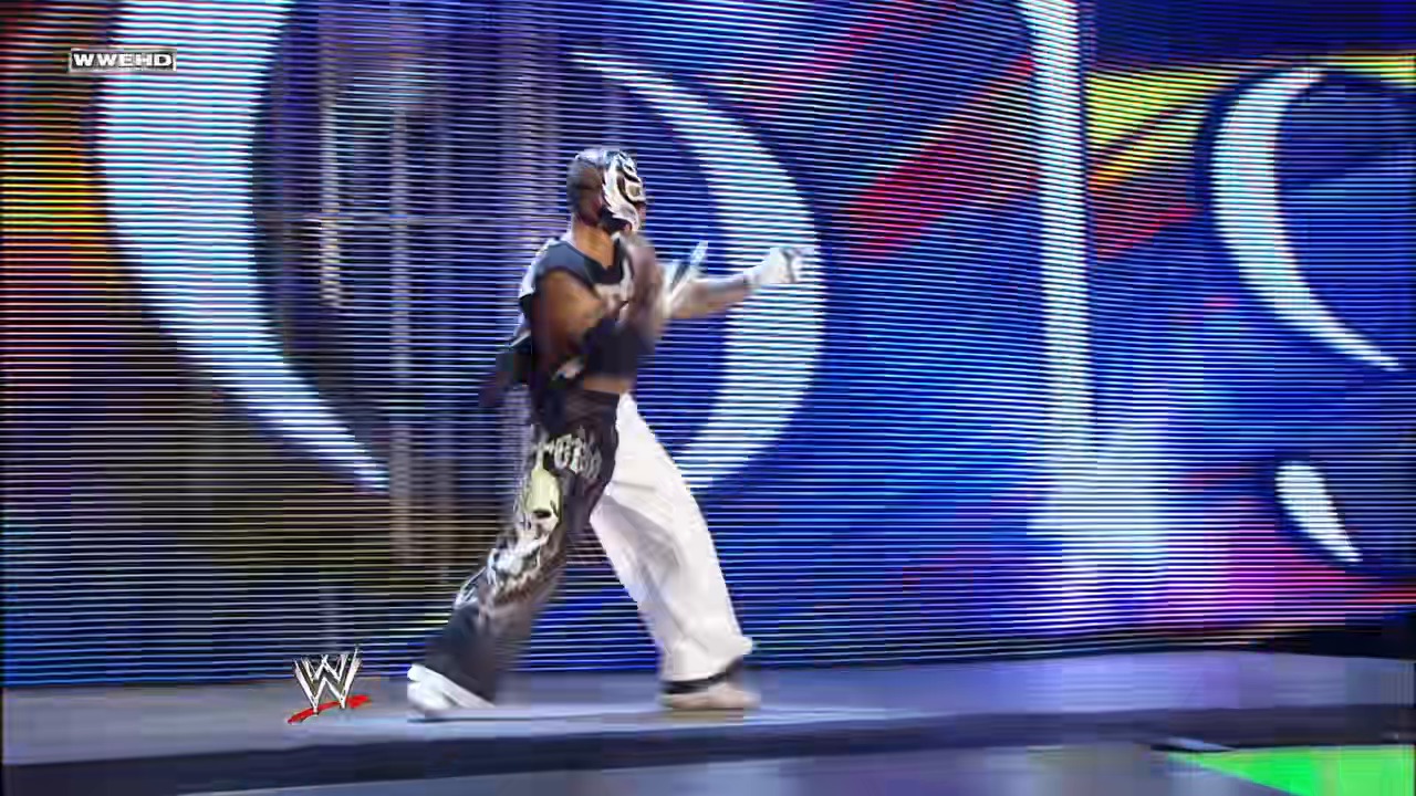 A frame from WWE Raw (January 12, 2009) at 13:33. A masked wrestler in black and white attire performs in the WWE ring, striking a dramatic pose with arms outstretched against a vibrant blue LED screen backdrop. The WWE logo is visible in the lower left corner of the broadcast.