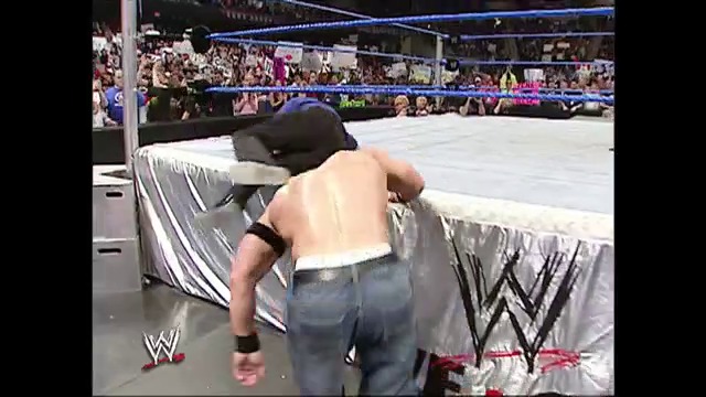 A frame from WWE SmackDown (February 18, 2005) at 49:42. Two shirtless wrestlers grapple near the ring ropes during a WWE SmackDown match, with a packed crowd visible in the background and the distinctive WWE logo and blue ring lighting visible in the arena.