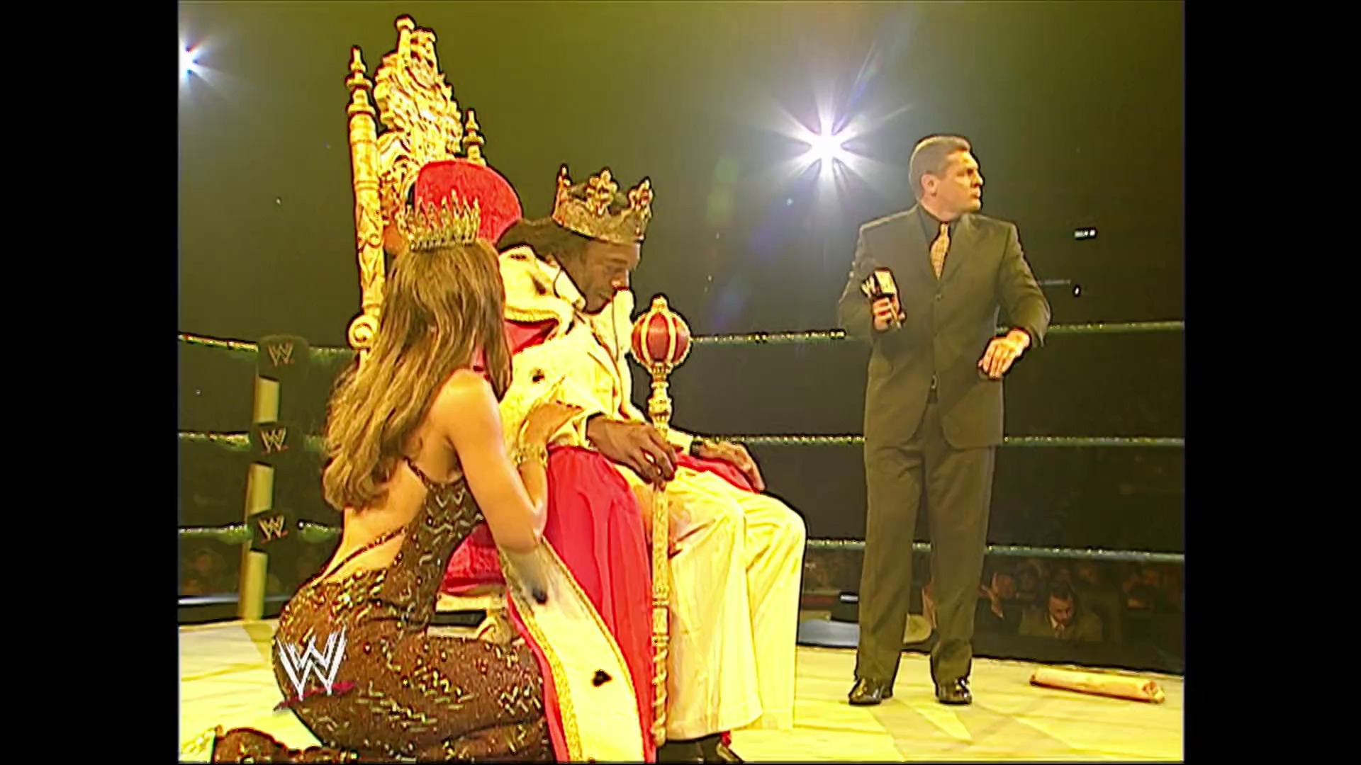 A frame from WWE SmackDown (July 14, 2006) at 4:57. A WWE wrestling scene featuring a performer seated on an ornate golden throne wearing a red and gold crown, with a woman in gold attire beside him and a commentator in a brown suit holding a microphone in the ring.