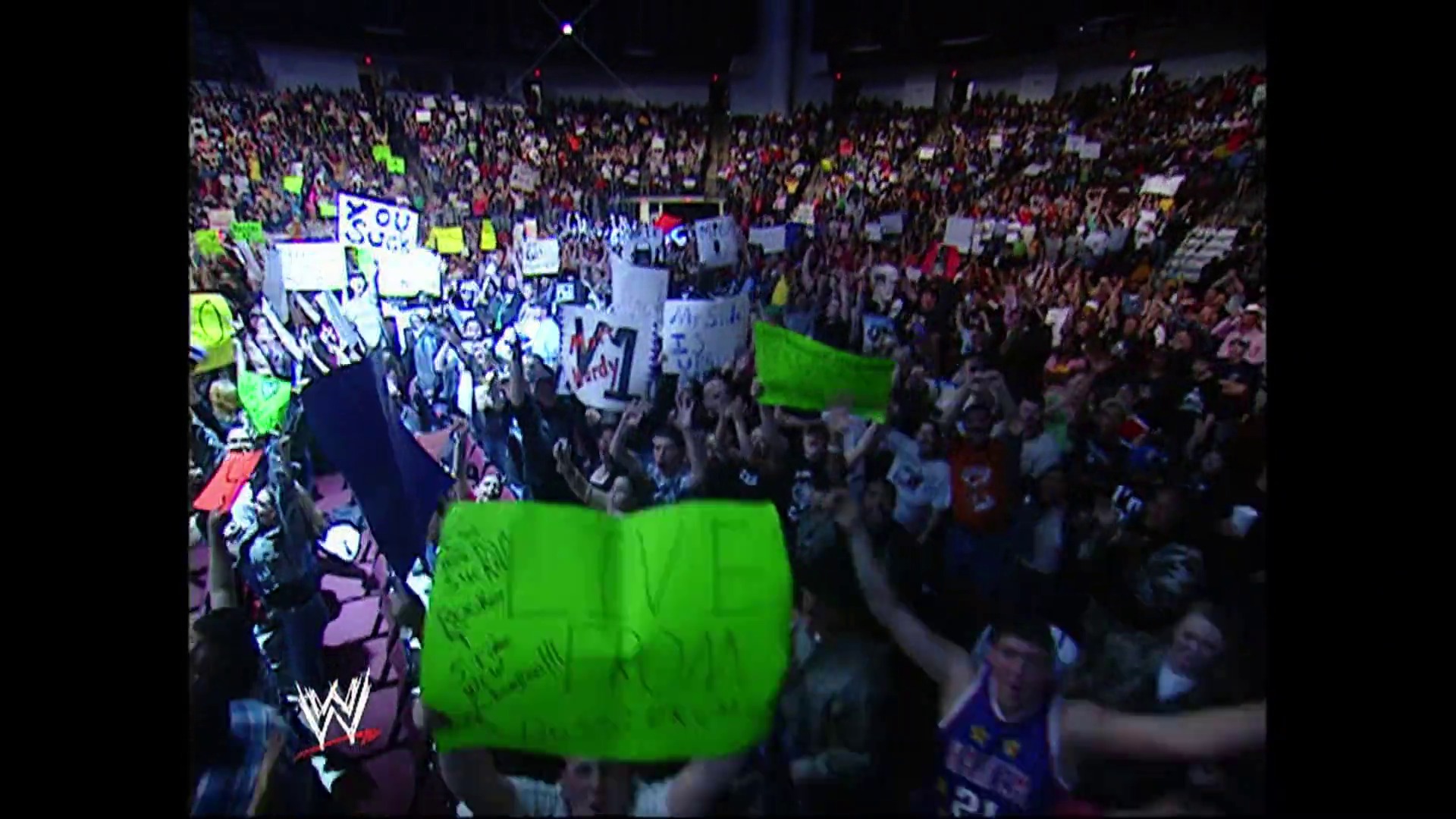 A frame from WWE SmackDown (March 17, 2006) at 2:15. A packed arena during a WWE SmackDown event features enthusiastic fans holding signs and cheering, with the WWE logo visible in the lower left corner. Green graphics and broadcast overlays are visible on screen during the live televised match coverage.