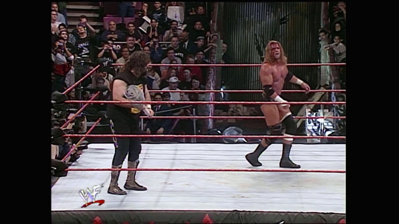 A frame from Royal Rumble (2000) at 1:22:20. Two wrestlers face off in a WWF ring with the crowd visible behind the ropes. One wrestler wears black attire while the other is shirtless, with the WWF logo visible on the mat.