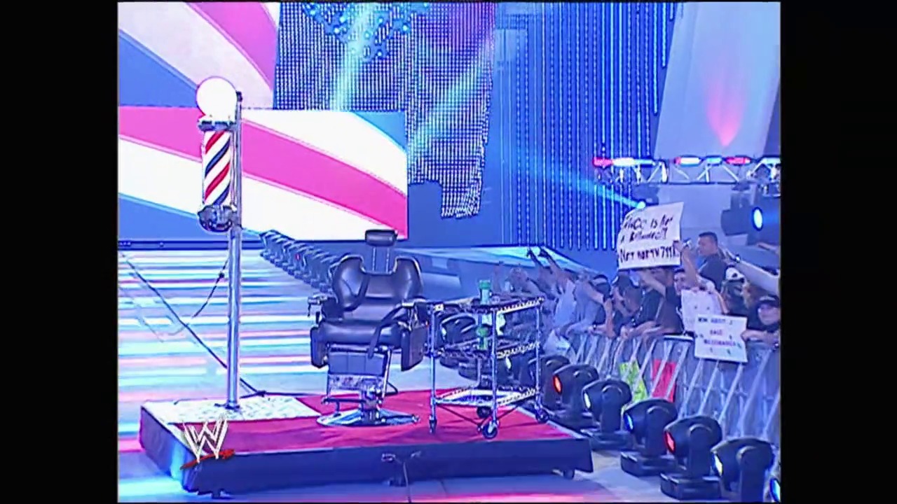 A frame from WrestleMania 23 (2007) at 2:06:48. A WWE wrestling ring setup featuring a barber pole and styling chair on the red mat, with the crowd visible behind barriers. Colorful blue and pink geometric graphics dominate the arena backdrop, characteristic of WrestleMania 23's vibrant production design.