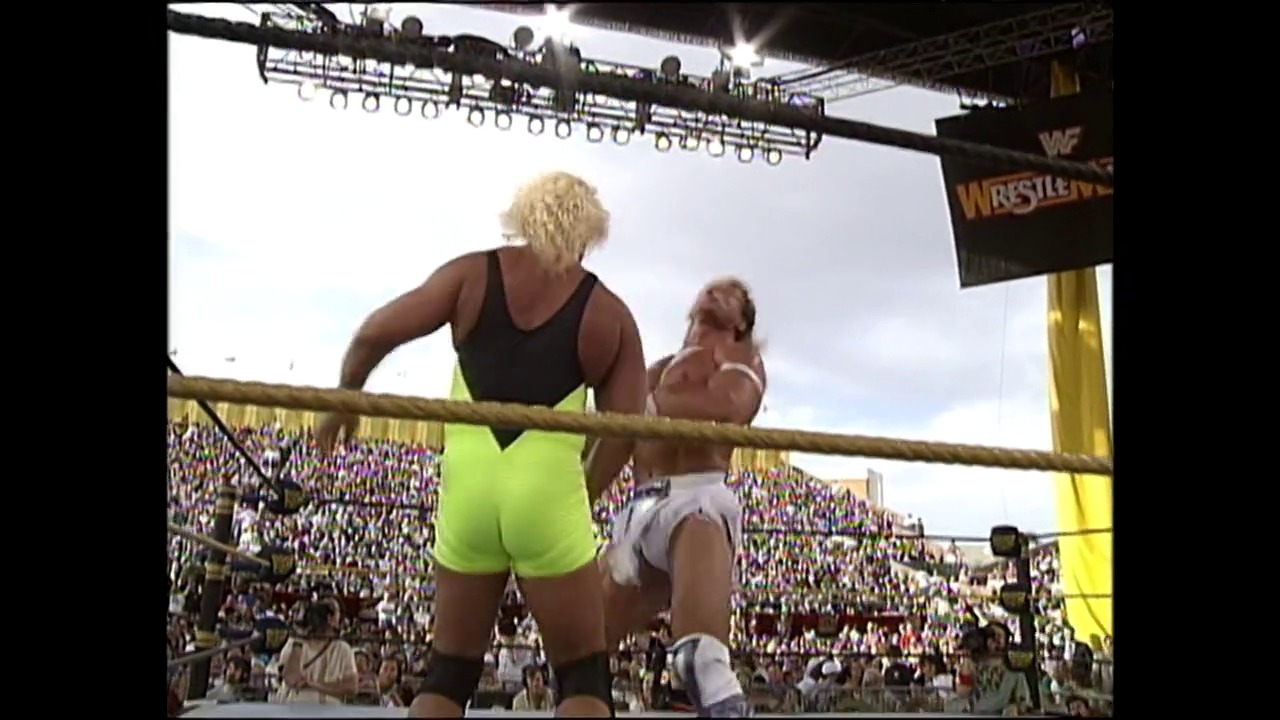 A frame from WrestleMania IX (1993) at 1:56:20. Two wrestlers face off in a WWF ring before a packed stadium crowd at WrestleMania IX. One wrestler wears bright neon yellow-green shorts while the other is shirtless, with the event's banner visible above the ring.