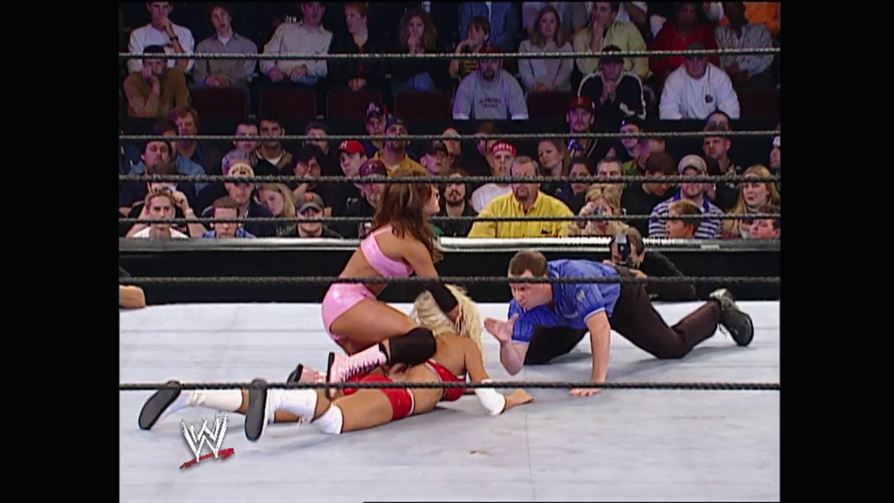 A frame from WWE No Mercy (2002) at 24:27. A WWE wrestler in pink attire dominates opponents in the ring while a full crowd watches from the stands. The white wrestling ring is surrounded by black ropes, with the WWE logo visible in the corner.