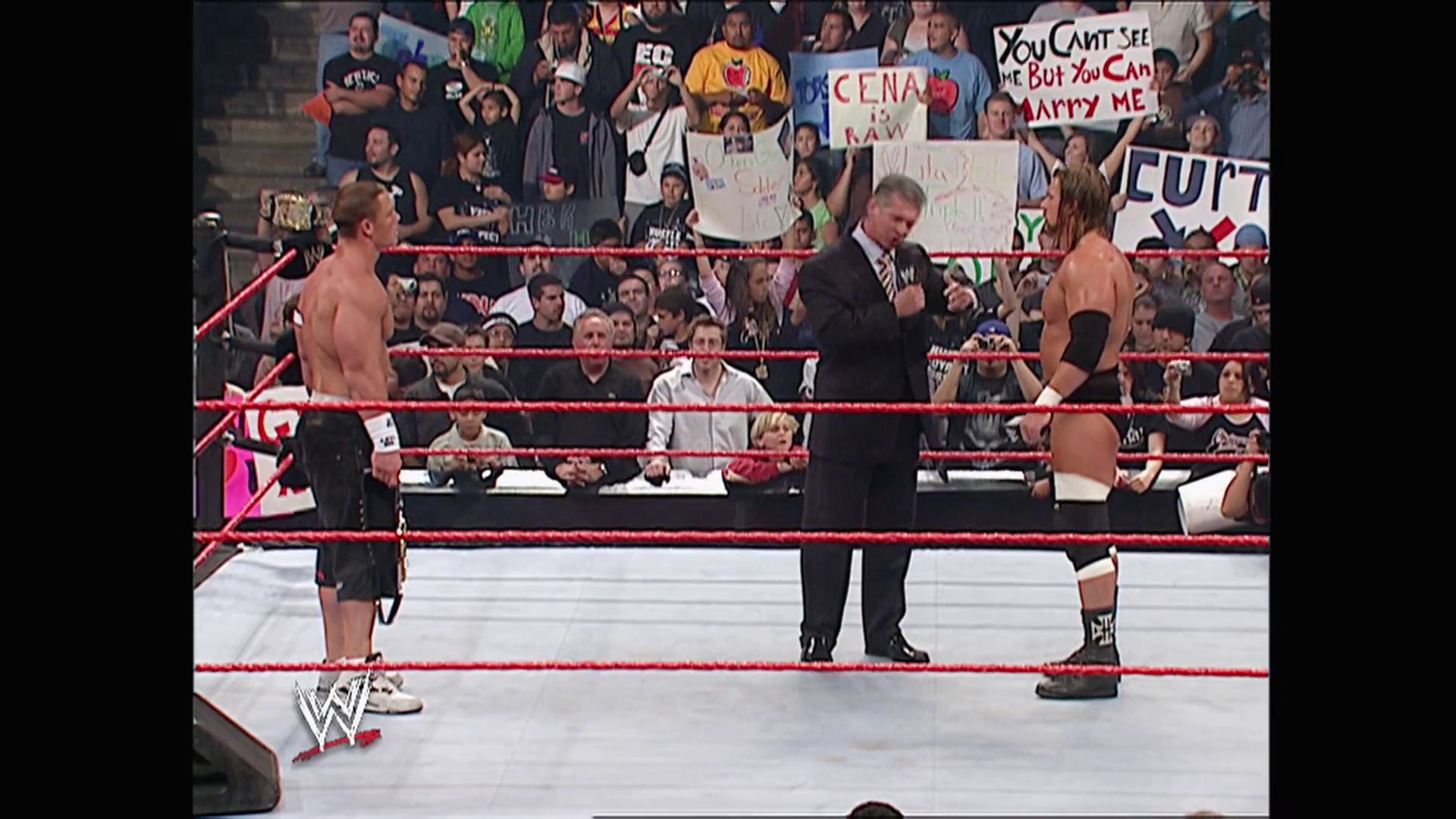 A frame from WWE Raw (May 8, 2006) at 12:48. A WWE Raw match in progress featuring two shirtless wrestlers in a red-roped ring with an official referee in a striped shirt between them. The crowd holds signs in the background, including one referencing "Cena," while the WWE logo is visible in the lower left corner.
