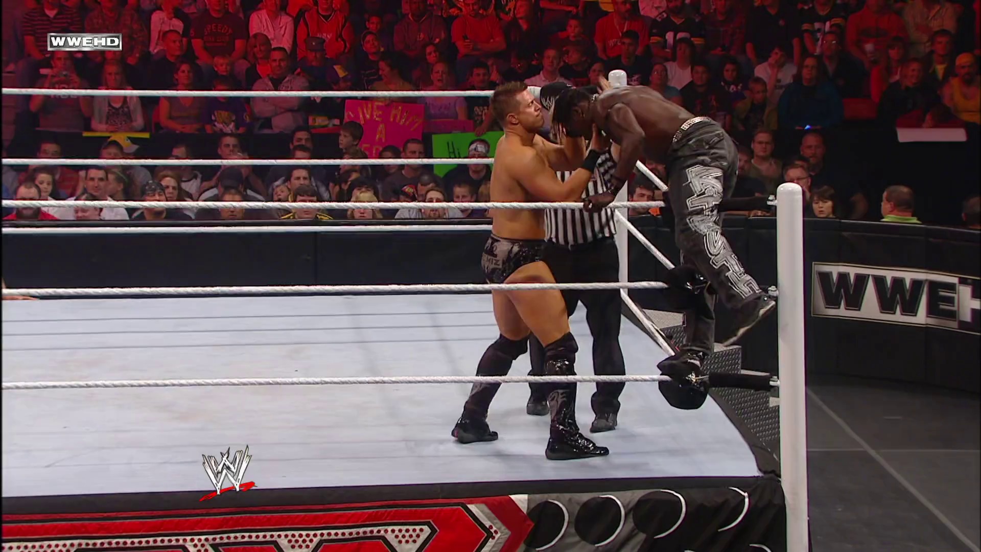 A frame from WWE Raw (October 25, 2010) at 53:49. Two wrestlers engage in combat inside a WWE ring, with one opponent pinned against the ropes while a referee monitors the action. The crowd fills the arena in the background under red lighting, with WWE branding visible on the ring apron and broadcast graphics.