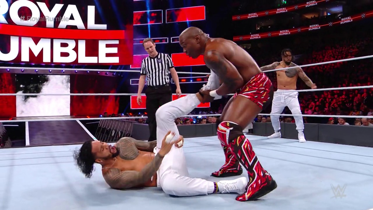 A frame from WWE Royal Rumble (2018) at 36:27. A wrestler in red and gold attire stands over a fallen opponent on the white WWE ring during Royal Rumble, with a referee in striped shirt nearby and cheering crowd visible in the background behind the branded barriers.