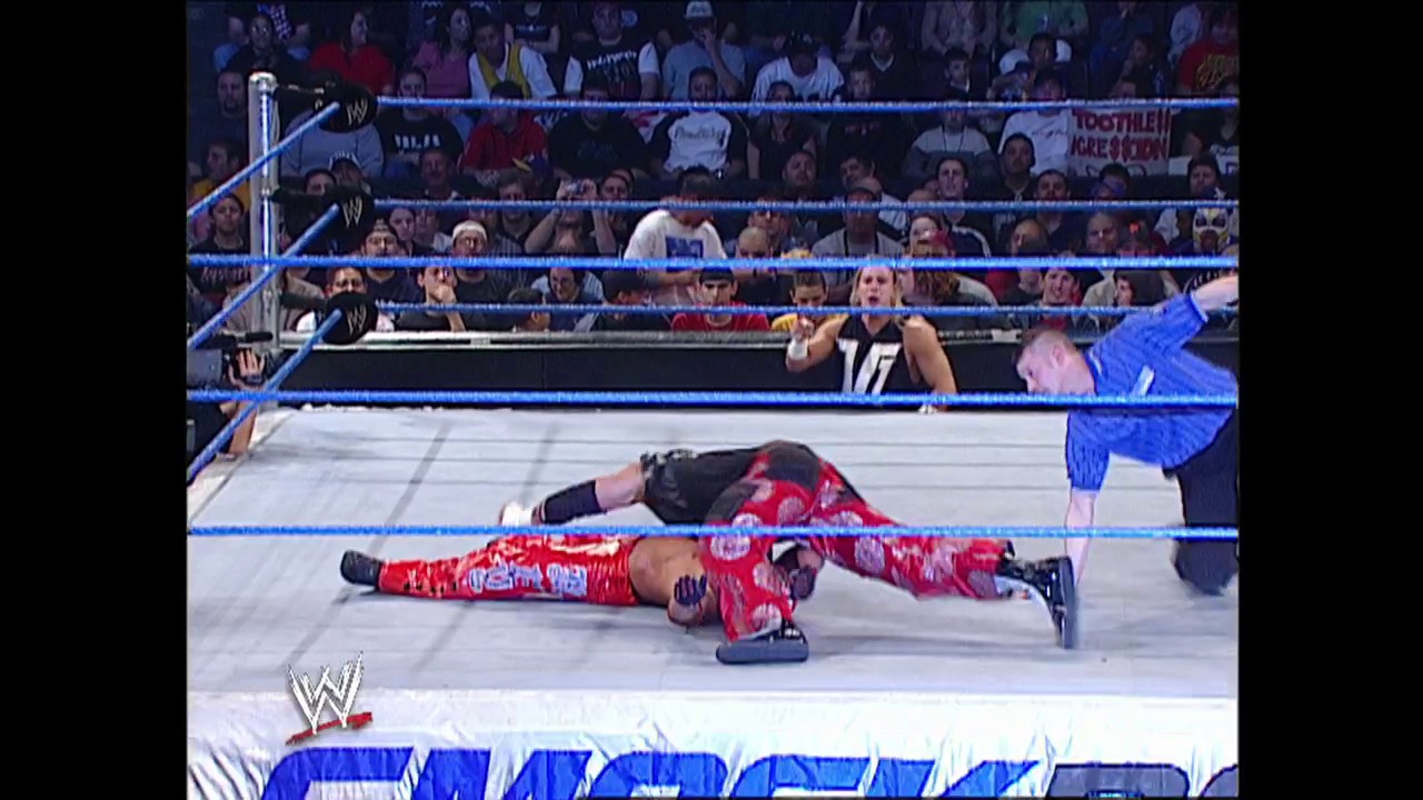 A frame from WWE SmackDown (February 13, 2003) at 23:29. A WWE wrestler in red and white attire lies on the canvas during a match, while an opponent in blue stands nearby. The crowd fills the arena seats in the background, with the blue-roped ring and WWE logo visible at the bottom of the frame.