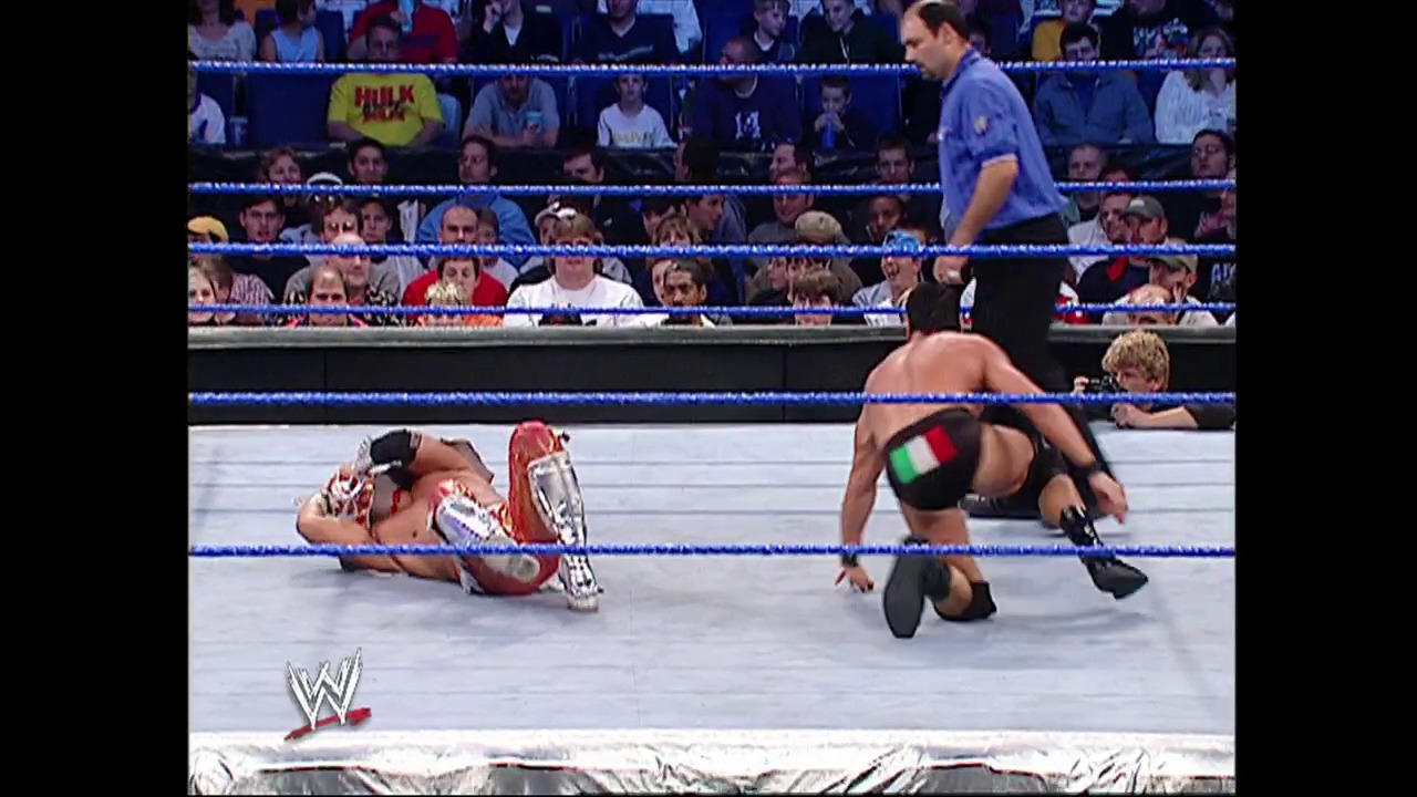 A frame from WWE SmackDown (November 6, 2003) at 59:52. Two wrestlers are engaged in action inside a WWE ring with blue ropes, with one opponent on the mat while the other remains standing. The crowd fills the arena stands in the background, and a referee oversees the match while the WWE logo is visible at the bottom of the frame.