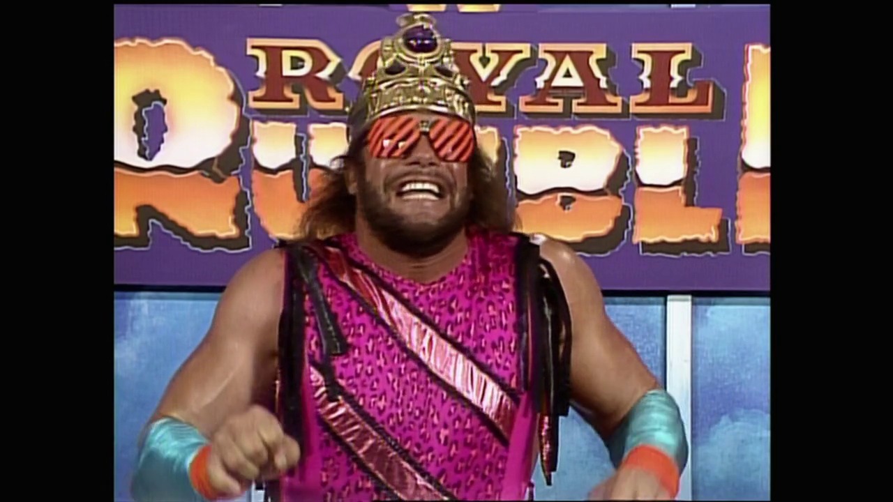 A frame from Royal Rumble (1990) at 1:33:10. A wrestler wearing pink and purple attire with striped sunglasses and a gold crown stands in front of the "Royal Rumble" logo backdrop, displaying an enthusiastic expression during the 1990 wrestling event.